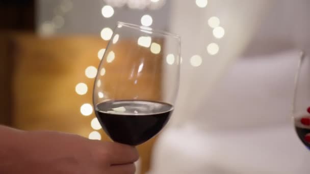 Primo piano di un uomo e una donna che tintinnano bicchieri di vino rosso contro una bella ghirlanda di Natale luminosa. Le persone innamorate celebrano la festa e battono i bicchieri in un'atmosfera calda e piacevole.. — Video Stock