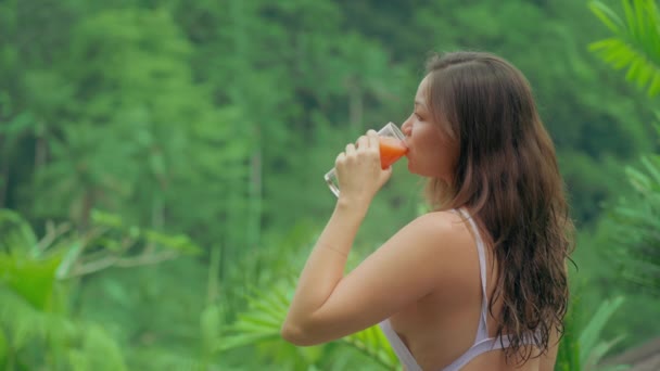 Una hermosa joven en traje de baño blanco bebe jugo de naranja recién exprimido sobre el fondo de una selva tropical verde con palmeras en un día soleado. Una mujer bebe jugo de un vaso. — Vídeos de Stock