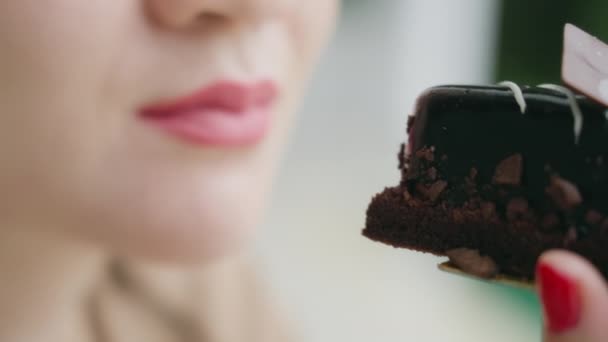 Zbliżenie dziewczyny z czerwonymi szminkowymi ustami gryzącej eklerkę pokrytą ciemną czekoladą. Kobieta z czerwonym manicure ma pyszny deser czekoladowy na rozmazanym biało-zielonym tle. Zwolniony ruch — Wideo stockowe
