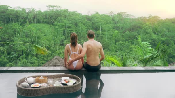 A couple in love watching the sunset sitting on the edge of the pool with a view of the tropical jungle and the sunset sky. A man in black shorts gently hugs a girl in a white swimsuit. — Stock Video