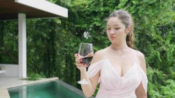 Uma linda garota sommelier em um vestido de noite rosa olha para um copo de vinho tinto, cheira seu cheiro. Uma menina asiática, conhecedora de vinhos, estuda uma nova variedade no fundo de uma piscina. — Vídeo de Stock