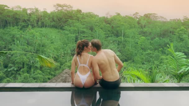 A couple in love, a man in black shorts and a girl in a white swimsuit, are sitting on the edge of the pool with a view of the rainforest and sunset. Male pushes the girl and she falls into the pool. — Stock Video