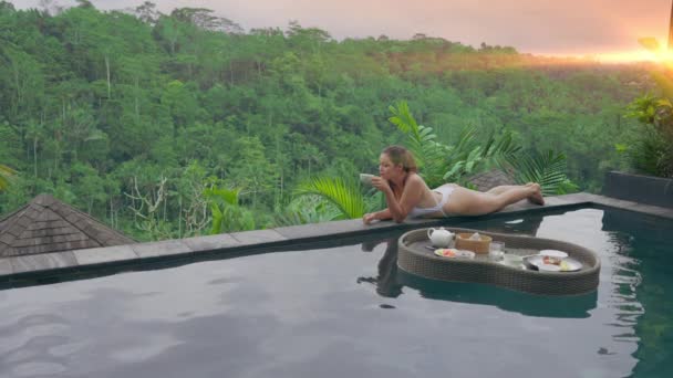 Piękna dziewczyna w białym stroju kąpielowym leży na skraju basenu z widokiem na tropikalny krajobraz w promieniach słońca. Kobieta opala się na brzegu basenu i pije kawę.. — Wideo stockowe