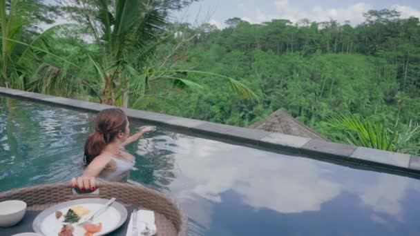 日の出に熱帯ジャングルを望む豪華なホテルスパでエキゾチックな料理を楽しむスイミングプールで朝食を持っている旅行女性。白い水着の女の子と浮動小数点の朝食。スローモーション. — ストック動画