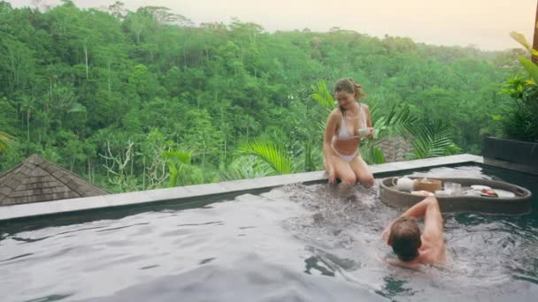 Una chica en traje de baño blanco y un chico se divierten chorros en la piscina de un hotel spa con vistas a la selva tropical en la luz del atardecer. Una pareja joven se divierten en la piscina con vista al bosque tropical — Vídeos de Stock