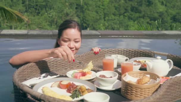 Siyah saçlı genç bir kız havuzda yüzüyor ve üzerinde yüzen bir kahvaltı olan bir sepetten ananas yiyor. Bir kadın, tropik bir ormana bakan, palmiyeli bir otelde dinleniyor.. — Stok video