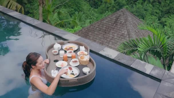Podróżniczka jedząca śniadanie w basenie, delektująca się egzotycznym jedzeniem w luksusowym hotelowym spa z widokiem na tropikalną dżunglę o wschodzie słońca. Dziewczyna w stroju kąpielowym je jedzenie z kosza z pływającym śniadaniem. — Wideo stockowe