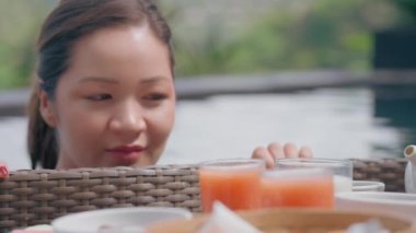 Aç bir kızın, üzerinde yüzen bir kahvaltı olan sepete bakışıyla. Genç ve güzel bir Asyalı kız seçkin bir otelin havuzunda duruyor ve güneşli bir günde yiyeceklere bakıyor..