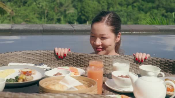 Beyaz mayo giymiş aç bir kız, üzerinde yüzen bir kahvaltı olan sepete bakıyor. Güzel bir kadın spa otelinin havuzunda güneşli bir günde ormanın karşısında yiyeceğe bakıyor.. — Stok video