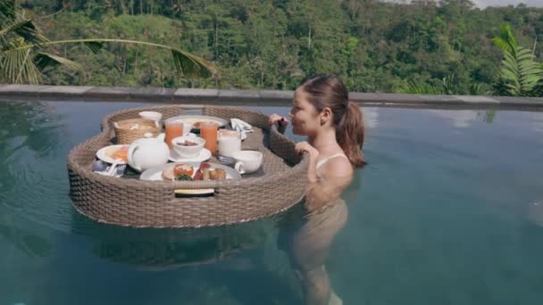 Piękna dziewczyna w białym stroju kąpielowym stoi w hotelowym basenie i odwraca się ze wspaniałym koszem pływającego śniadania. Atrakcyjna Azjatka relaksuje się w basenie ze wspaniałym widokiem. — Wideo stockowe