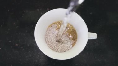 Bir insan sabah kahvesini kaynayan suyla yavaş çekimde demler. Siyah bir masanın üzerinde duran beyaz bardağın üst görüntüsü sıcak su akışının akmaya başladığı yerde..