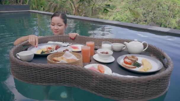 一个漂亮的亚洲女孩在游泳池里带着漂浮的早餐。吃煎蛋卷的女人一个女孩正在一个绿色的背景下，从一个漂浮的篮子里在游泳池里吃早餐 — 图库视频影像