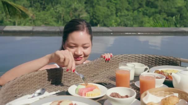 Havuzdaki güzel bir kız kahvaltıdan çatalla ananas almaya çalışıyor, çatalı kayıyor ve tatlı tatlı gülümsüyor. Lüks bir spa otelinde tatilde olan Asyalı bir kız. Yavaş çekim. — Stok video