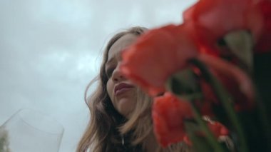 Uzun saçlı, parlak gözlü, kırmızı çiçekler ve bulutlu bir gökyüzüne karşı bardak tutan üzgün bir kızın yakın çekimi. Başarısız bir randevu ve gerçekleşmemiş bir rüyadan ağlayan bir kadın.