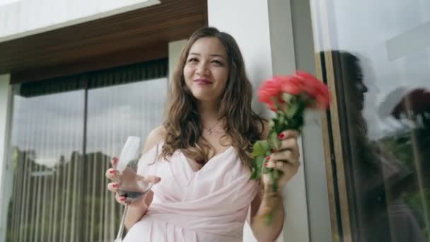 Ein zufriedenes Mädchen in einem rosafarbenen Kleid mit lockeren dunklen Haaren steht mit einem Strauß roter Rosen und einem Glas Wein vor einer weißen Wand mit Fenstern. Asiatisches Mädchen lächelt und winkt mit einem Strauß Rosen. — Stockvideo