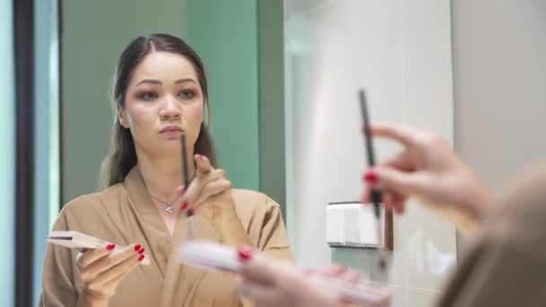 Красива дівчина з чорним волоссям у бежевій халаті стоїть біля дзеркала у ванній кімнаті. Азійська дівчина з червоною манікюр блимає її очі, стоячи перед дзеркалом.. — стокове відео