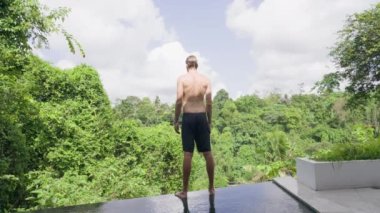 Siyah şortlu uzun boylu atletik bir genç havuzun kenarında duruyor ve sabah egzersizleri yapıyor. Güzel bir sırtı olan genç bir adam, tropikal bir manzaranın arka planına yaslanır..