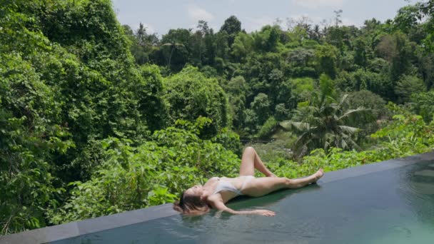Una hermosa chica delgada en un traje de baño blanco está tomando el sol en el lado de una piscina de lujo con vistas a la selva tropical en un día soleado. Un modelo superior con el pelo largo es relajante en un hotel spa o villa. — Vídeos de Stock