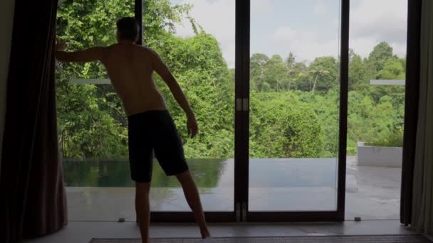 De manhã, um jovem bem sucedido abre as cortinas e dois saem e saltam para a piscina com vista para a floresta tropical e o céu azul com nuvens. Um homem rico começa a nadar num luxo — Vídeo de Stock