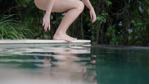 Uma menina bonita em um maiô branco senta-se ao lado de uma piscina branca moderna e acaricia a água de cor esmeralda com o brilho do sol. Uma menina viajante de férias em um dia ensolarado. — Vídeo de Stock
