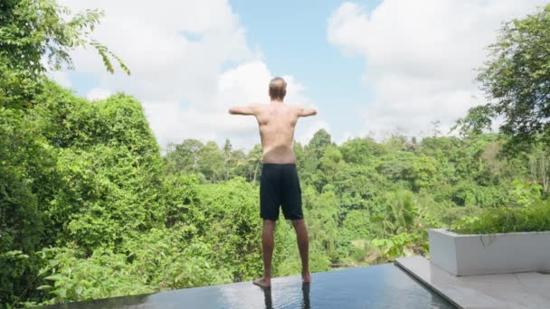 Wysoki wysportowany młodzieniec w czarnych spodenkach stoi na krawędzi basenu i wykonuje poranne ćwiczenia. Młody człowiek z pięknym kręgosłupem trenuje na tle tropikalnego krajobrazu.. — Wideo stockowe
