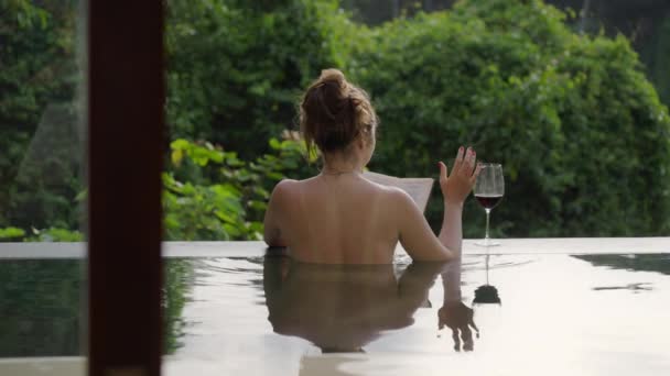 Dziewczyna siedzi w luksusowym basenie widokowym czytając książkę i pijąc wino na tle zielonych drzew w ciepłym świetle zachodu słońca. Kobieta ze śladami oparzeń słonecznych spoczywa w hotelu spa.. — Wideo stockowe