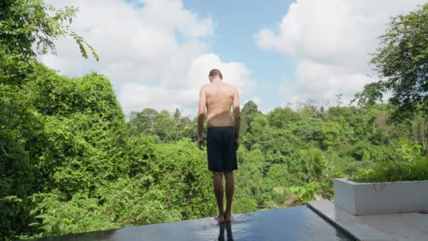 Високий атлетичний молодий чоловік в чорних шортах стоїть на краю басейну, роблячи йогу. Чоловік тренується на тлі тропічного пейзажу і блакитного неба з білими хмарами. повільний рух . — стокове відео