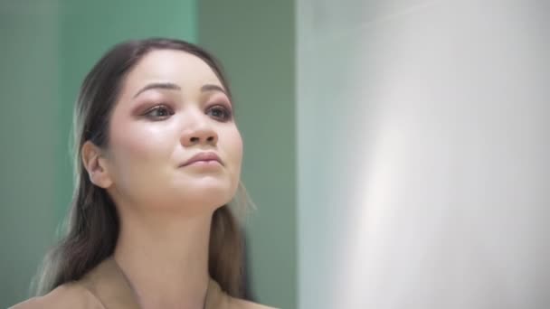 A beautiful contented girl with long dark hair looks in the mirror and examines her face and rejoices in her beauty. Asian girl looks at her face after spa treatments in the bathroom. — Stock Video