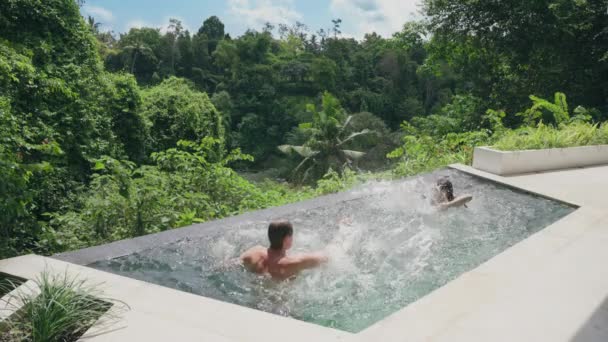 Dziewczyna w białym kostiumie kąpielowym i facet zabawiają się tryska w basenie hotelu spa z widokiem na las deszczowy w świetle zachodu słońca. Młoda para bawi się na basenie z widokiem na las tropikalny — Wideo stockowe