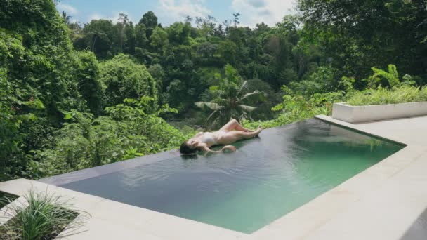 Um jovem caucasiano salta para a piscina e espirra uma menina tomando banho de sol durante o dia. Um casal de férias em uma piscina moderna vista de luxo divirta-se derramando água e espirrando rindo. Movimento lento. — Vídeo de Stock