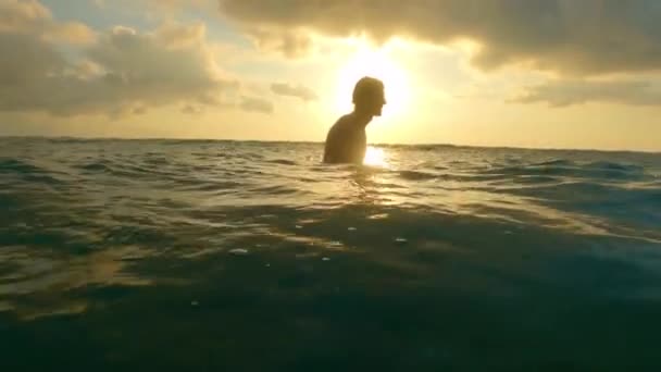 Un joven surfista guapo está sentado en una tabla en el océano y esperando una ola mientras disfruta de la puesta de sol. La silueta de un surfista caucásico sobre el fondo de un cielo con nubes y sol. — Vídeos de Stock