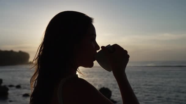 夕日の海を背景にマグカップから長髪の少女のシルエットが飲み物を飲んでいます。太陽の光に包まれた大きなマグカップから女の子がお茶を飲んでいます。スローモーション. — ストック動画