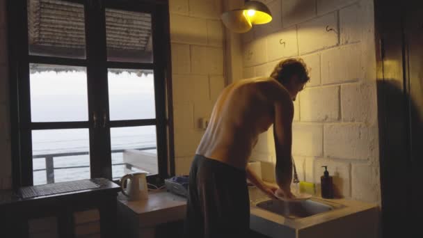 Przystojny wysoki surfer w spodenkach wchodzi do kuchni zapala światło i zaczyna zmywać naczynia. Sportowy atleta Ocean i salony są widoczne w świetle zachodu słońca w oknie. — Wideo stockowe
