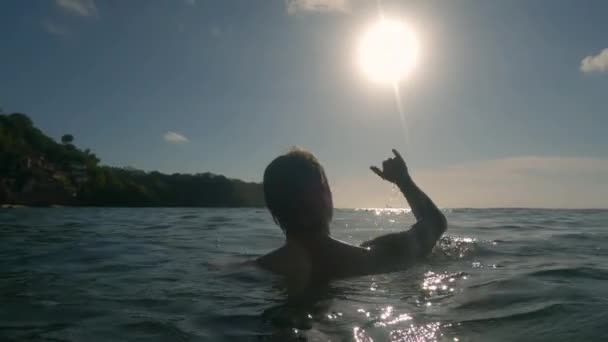 La silueta de un joven que muestra el gesto del surfista Shaka hacia el cielo sobre el fondo de las olas, el sol y el cielo azul. Un joven surfista nada en el agua y muestra un gesto de saludo de surfista. — Vídeos de Stock
