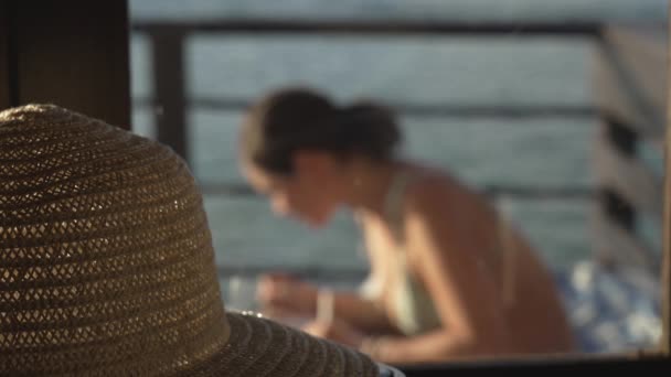 Großaufnahme eines niedlichen Strohhutes auf dem Hintergrund eines Künstlermädchens, das an einem sonnigen Tag ein Bild auf einem Seebrücke malt. Die Kopfbedeckung einer Schriftstellerin, die Reisenotizen in ihr Tagebuch schreibt. sonniger Tag. — Stockvideo