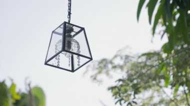 Rüzgarda savrulan kristal küreli güzel bir fener mavi gökyüzü ve yeşil ağaç dallarının arka planında sallanıyor. Açık hava tropikal bir restoranda avizenin kavramsal yakın çekimi. 4k