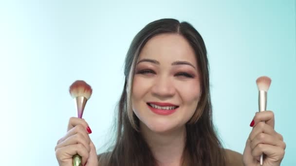 A close-up of a makeup artist girl who smiles and dances, holding makeup brushes in her hands and waving them as if they were maracas. Wonderful asian cheerful girl having fun and looking at camera — Stock Video