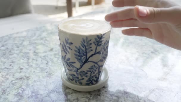 Κοντινό πλάνο ενός ανθρώπου με ένα ροζ μανικιούρ που παίρνει μια λευκή κούπα καφέ από ένα μαρμάρινο τραπέζι εστιατορίου μια ηλιόλουστη μέρα. Το κορίτσι παίρνει μια λευκή κούπα καφέ με ένα μπλε μοτίβο στο στυλ του Gzhel. — Αρχείο Βίντεο