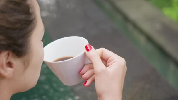 Közelkép egy piros manikűrös lányról, aki fehér pohárból issza a kávét egy medence vagy szökőkút hátterében vízzel. Lány iszik reggeli kávé háttér medence a szállodában. — Stock videók