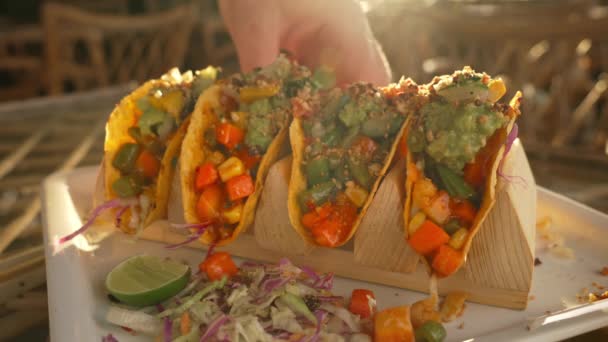 En person tager en appetitvækkende mexicanske tacos fra en hvid plade i stråler af varmt sollys. Middag med tacos med en lys fyldning og en drink, på et glasbord under solnedgang i en cafe. Nærbillede. – Stock-video