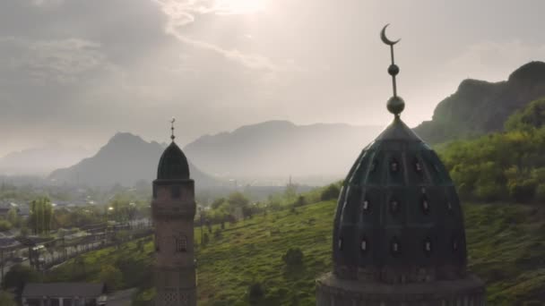 Повітряний політ між куполами мечеті на тлі міста, туманна зелена долина і великі гори в променях контурного сонця. Красивий пейзаж з приголомшливим — стокове відео