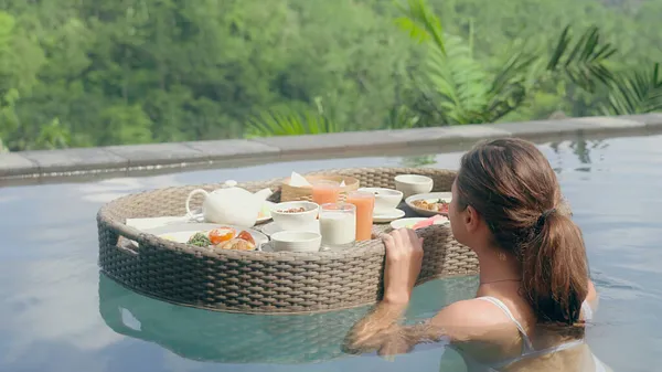 Ein schönes Mädchen im weißen Badeanzug mit roter Maniküre und schwimmendem Frühstückskorb schwimmt im Pool. Ein Mädchen und ein luxuriöses schwimmendes Frühstück vor dem Hintergrund des grünen Dschungels. — Stockfoto