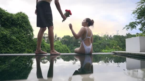 En flicka i vit baddräkt sitter på sidan av utsiktspoolen, killen i shorts närmar sig henne och ger henne bukett röda rosor. Mannen ger sin älskade flicka blommor, sätter sig bredvid henne och kramar henne. — Stockvideo