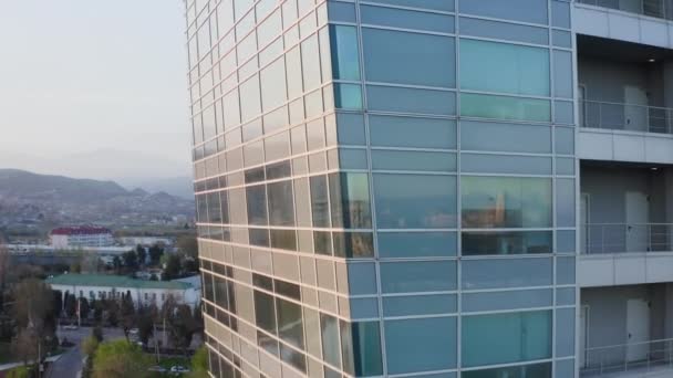 Luftaufnahme eines vorbeifliegenden Gebäudes mit Glasfenstern, in denen sich die Sonne spiegelt. Bauen in einer städtischen Umgebung, eine Moschee und große Berge sind in der Ferne sichtbar. Ein modernes Gebäude — Stockvideo