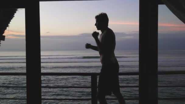 Silhuett av en sportig atletisk man boxning och träning stansar på balkongen mot bakgrund efter solnedgången kväll himlen. En idrottsman i shorts tränar flyganfall, skuggstrider. Långsamma rörelser. — Stockvideo