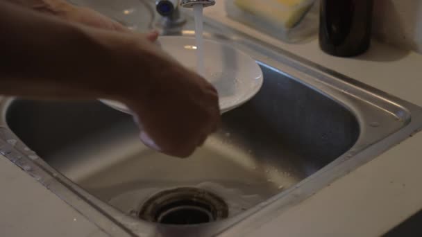 Personen sätter på vattnet och tvättar en vit tallrik med en svamp i ett järnfat i köket. Närbild. Människan klämmer rengöringsmedel på plattan och tvättar den i strålar av varmt artificiellt ljus. slow motion. — Stockvideo