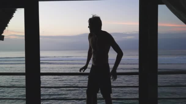 Красивый высокий мужчина в отпуске сидит на перилах обзорного балкона и наслаждается прекрасным пейзажем закатного неба. Молодой серфер смотрит на вечерний океан с красивым небом и облаками. Медленное движение. — стоковое видео