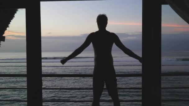 Egy gyönyörű, magas sziluett egy férfiról, aki felemeli a karját, és izmokat mutat az óceán és a naplemente égbolt hátterében. Egy sportos férfi edzés után egy bungaló erkélyén mutatja a bicepszét.. — Stock videók