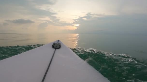 POV shot van een surfer op een witte surfplank peddelend in de horizon met de ondergaande zon. Een surfer zwemt op een plank in de oceaan door kleine golven met spatten water. — Stockvideo