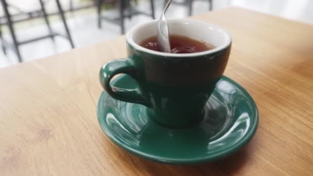 Dolly shot. A person stirs fragrant black tea with a spoon in a beautiful green mug standing on a wooden table in a bright cafe. English morning tea in a cafe with sunlight. Slow motion. — Vídeo de Stock